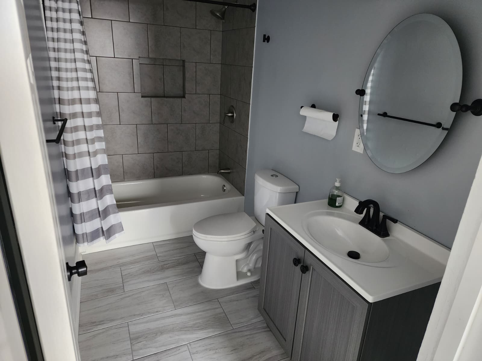 Finished bathroom with gray tiled tub surround, gray plank floor tile, white toilet and vanity with oval mirror.