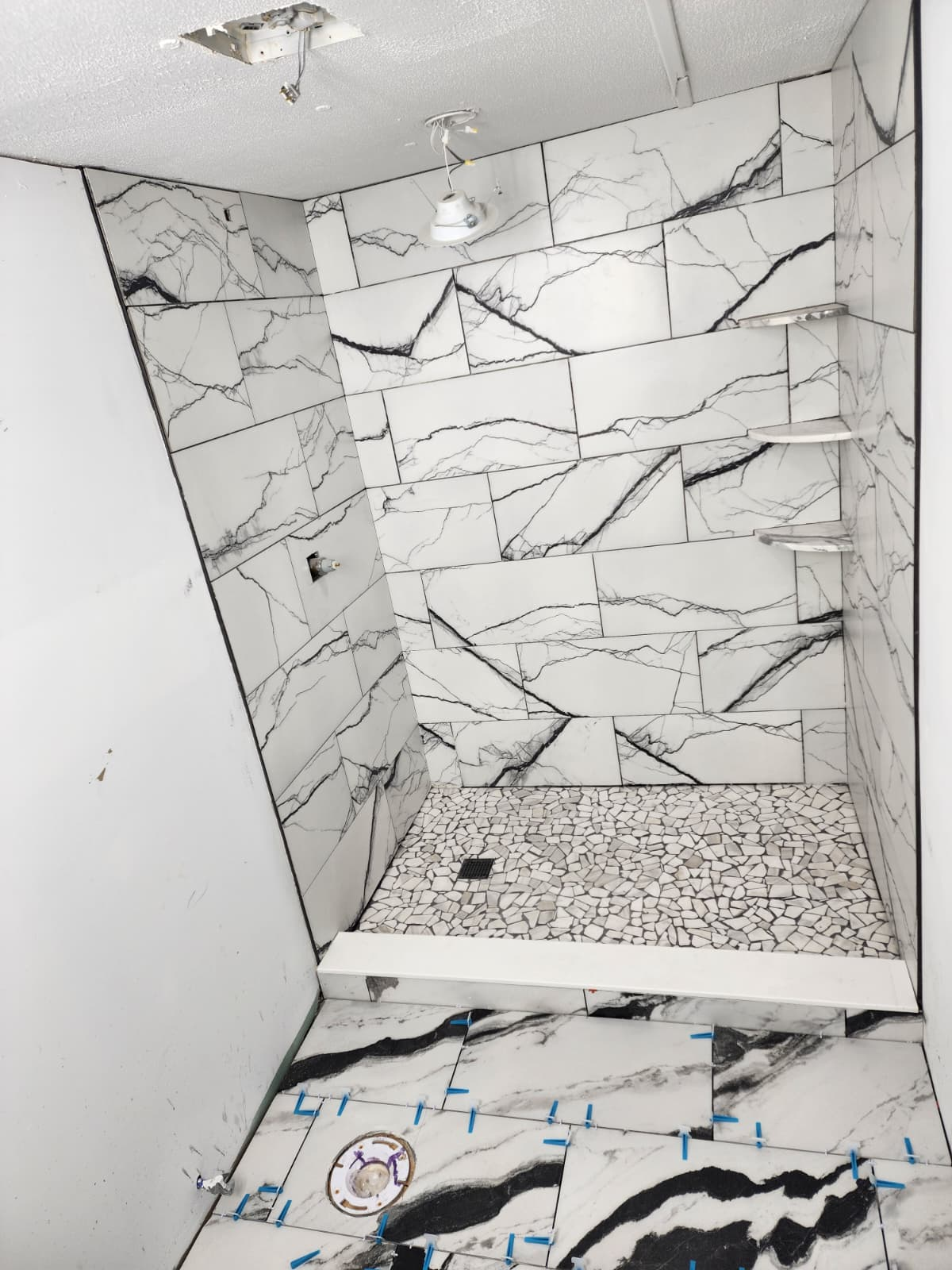Finished white marble-look tile shower with pebble mosaic floor and corner shelves.