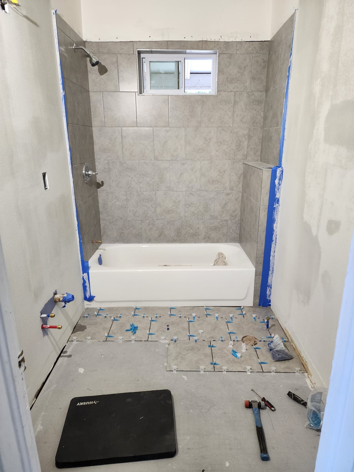 Bathroom tub surround tiled in gray with painter's tape at the edges and floor tiles in progress with leveling clips.