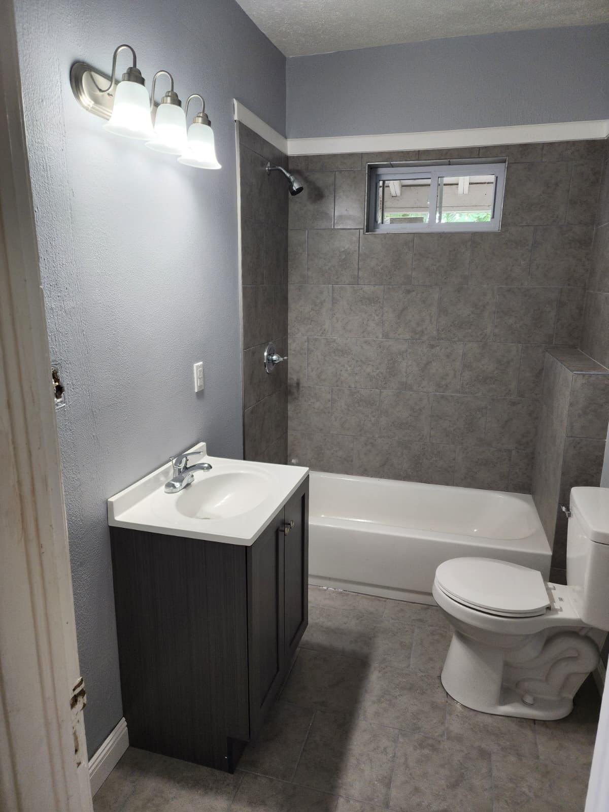 Completed bathroom with gray tiled tub surround, gray floor tile, blue-gray walls, vanity, toilet and vanity lights.