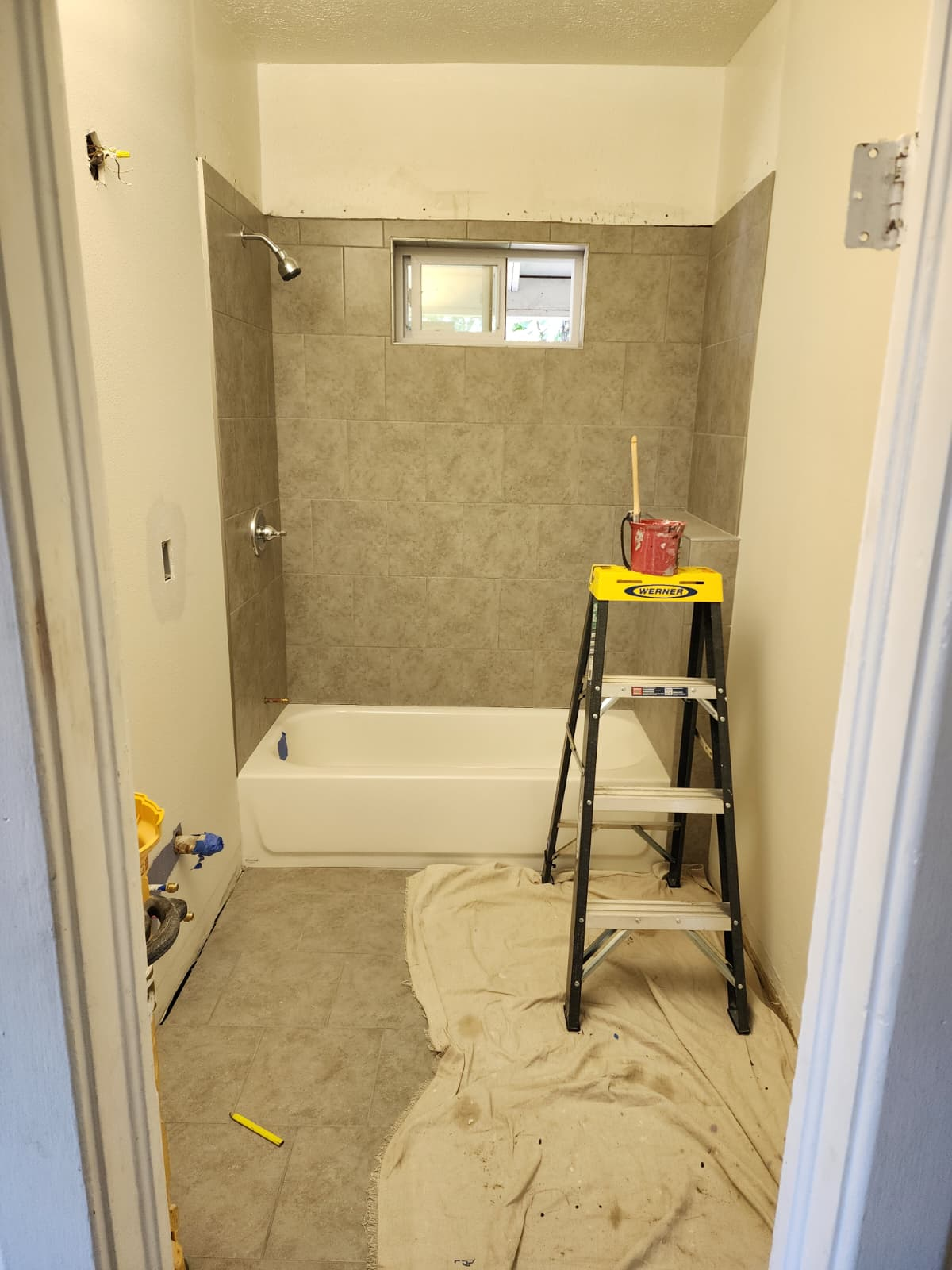 Bathroom remodel in progress with gray tiled tub surround, tiled floor, and a step ladder on a drop cloth.